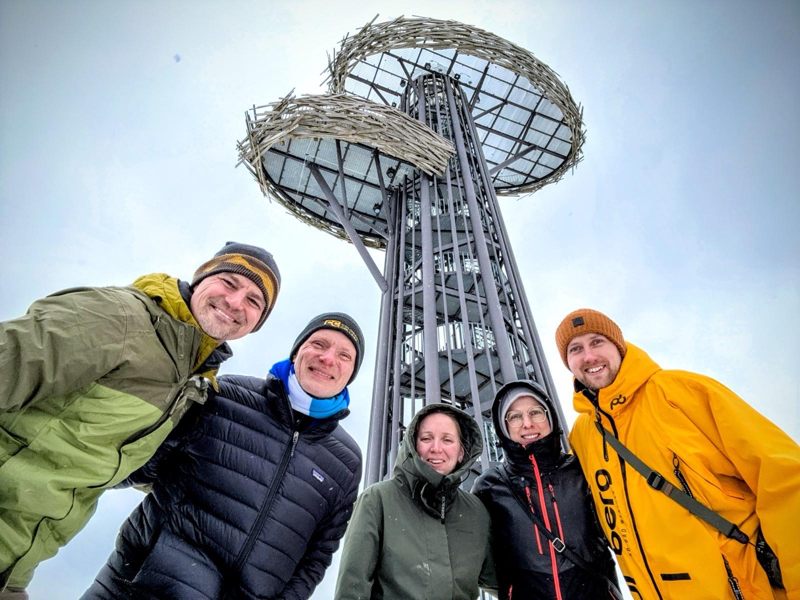 Five people gathered under a wooden viewing tower in falling snow